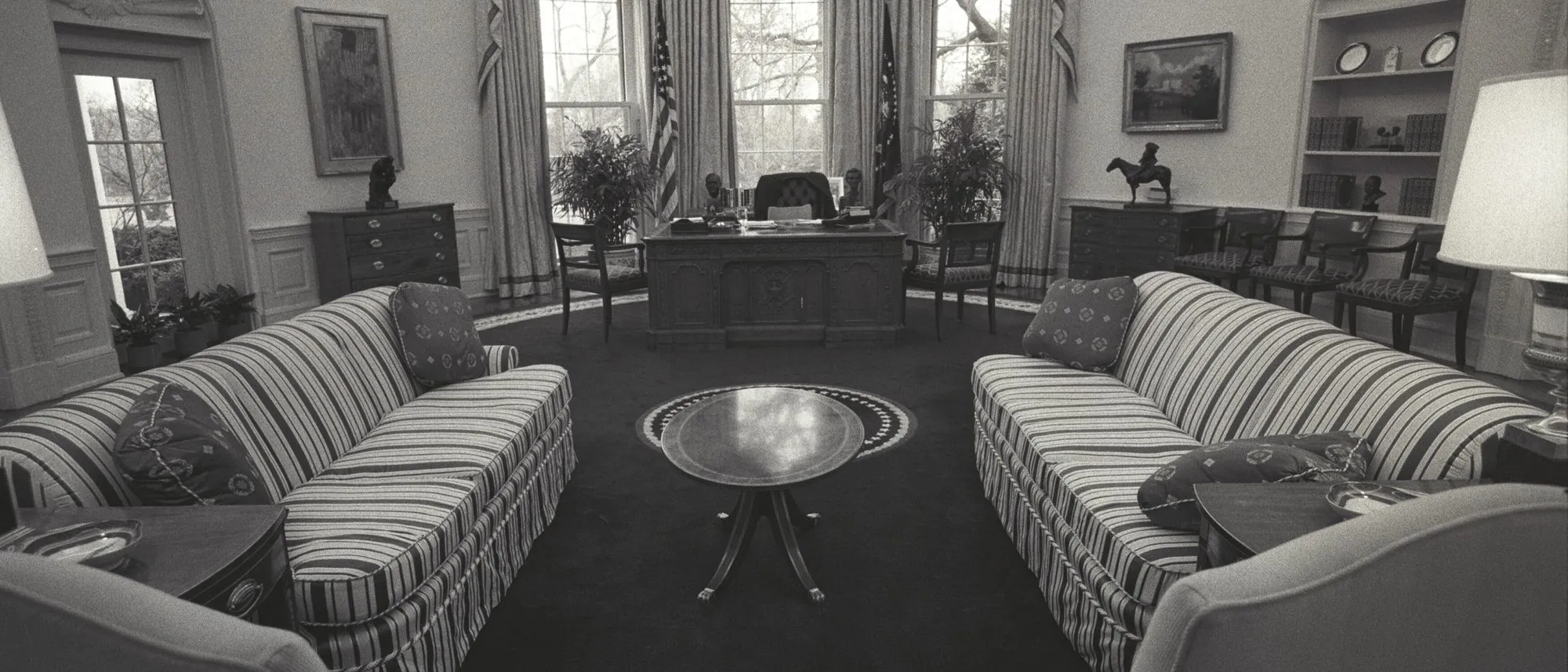 The Oval Office empty, photographed in black and white: the desk before the windows, two striped sofas facing each other and no one in the room