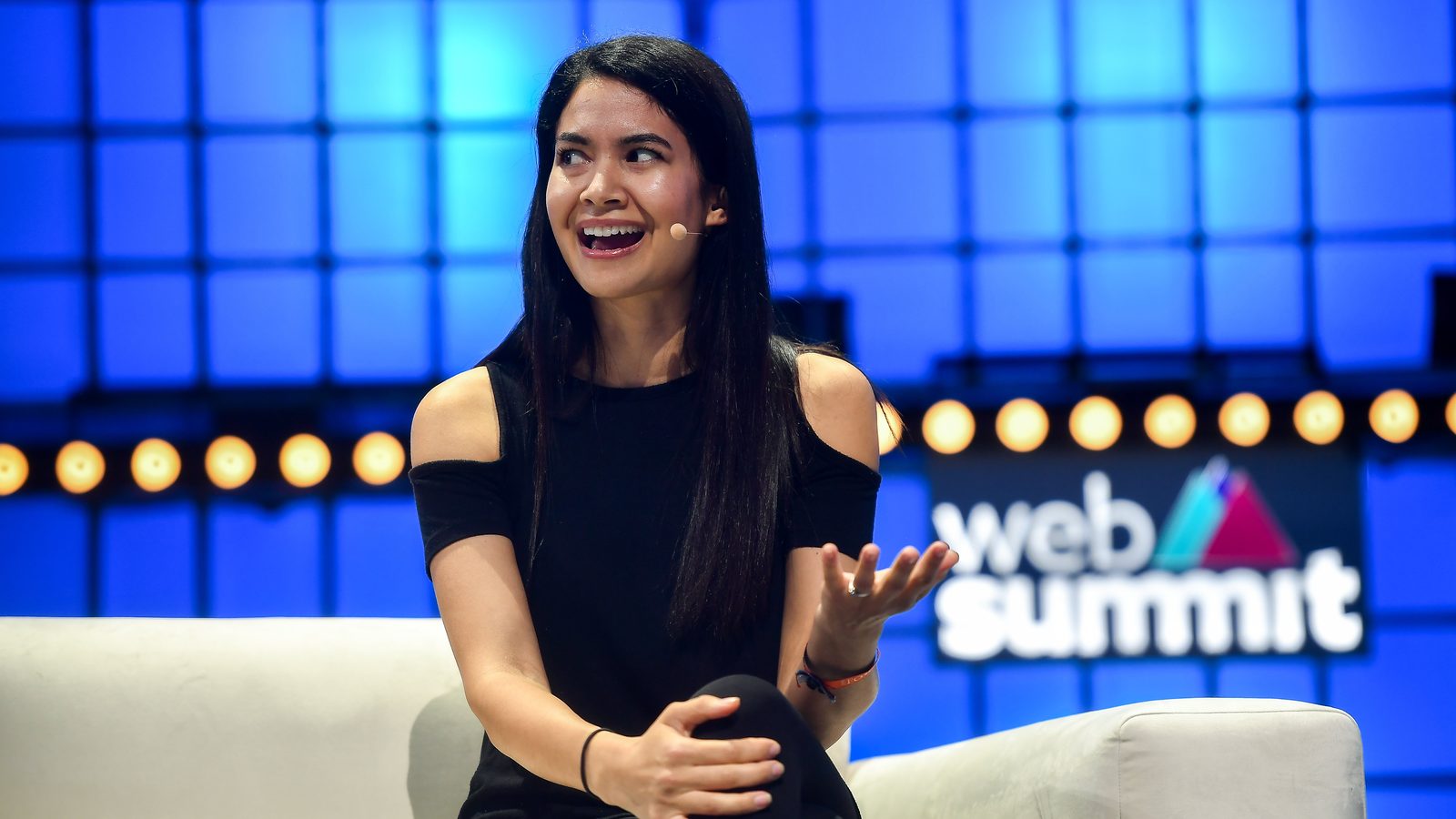 Melanie Perkins speaking from a white armchair on a stage lit in blue