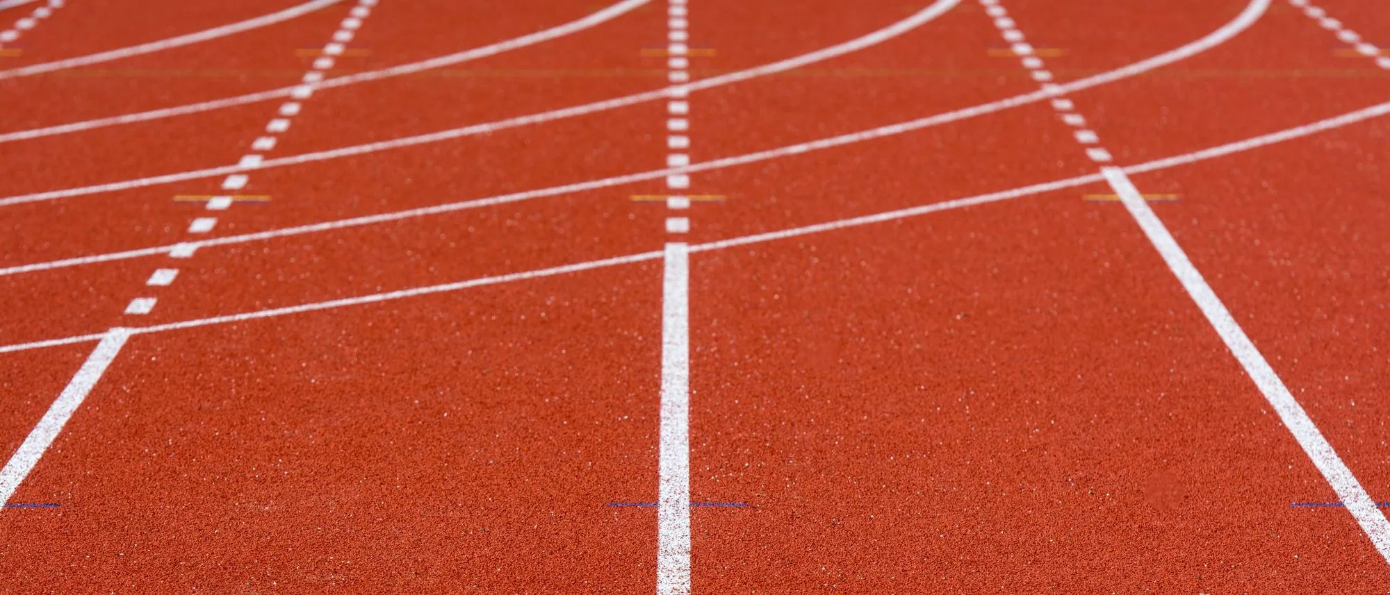 The red surface of an empty running track with white lane lines curving away.