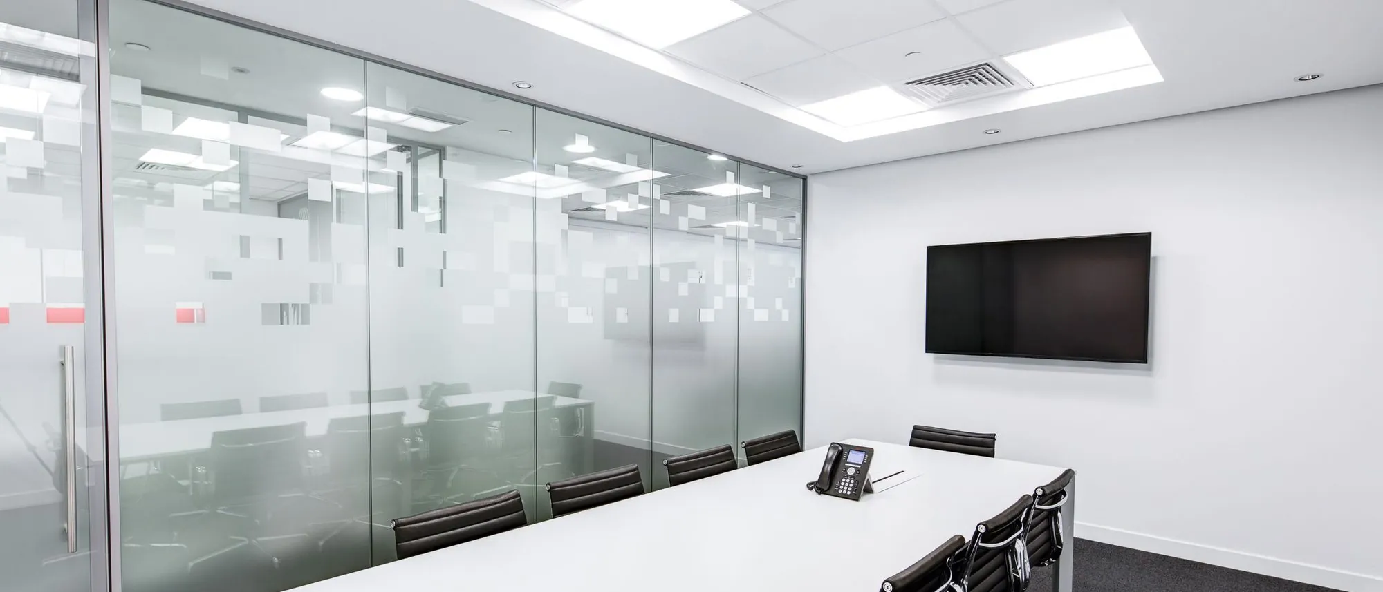 An empty glass-walled meeting room with a long white table and office chairs.
