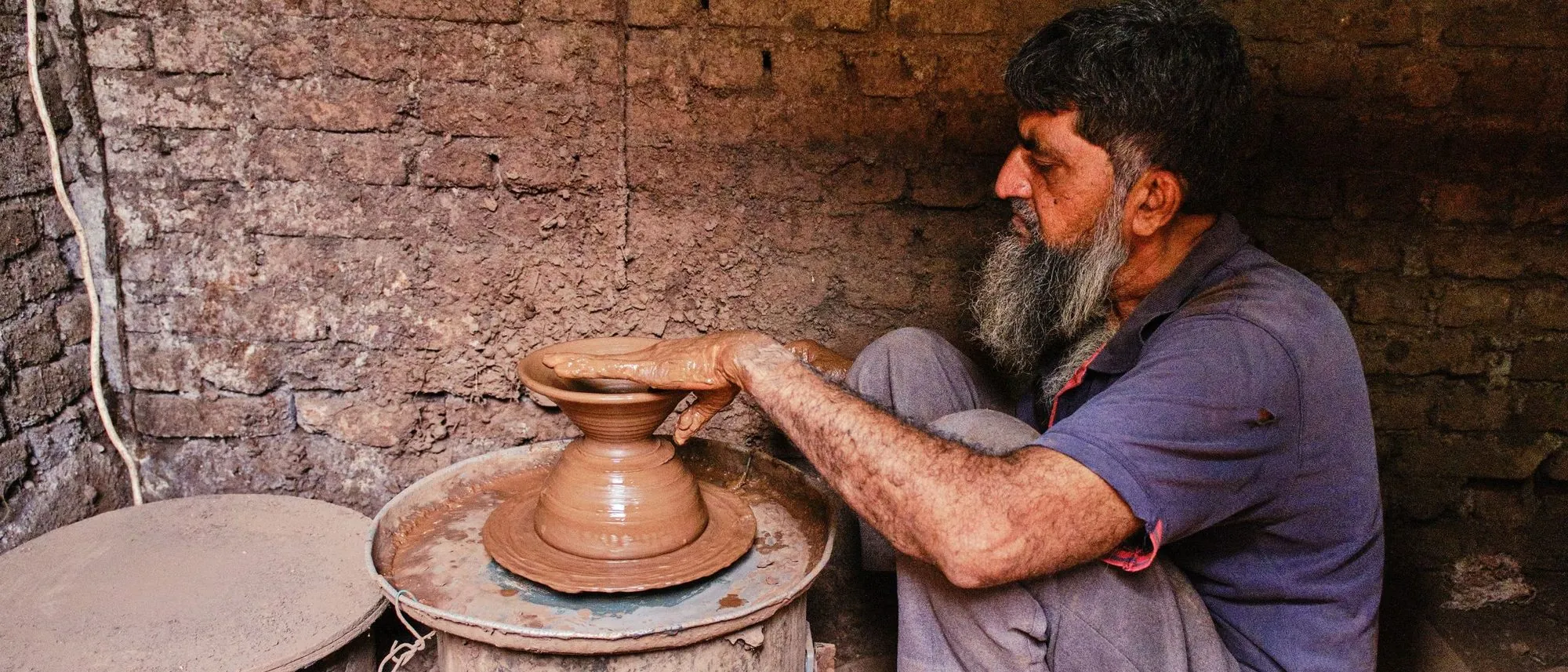 A potter at his wheel alone in a brick-walled workshop, shaping a clay pot by hand