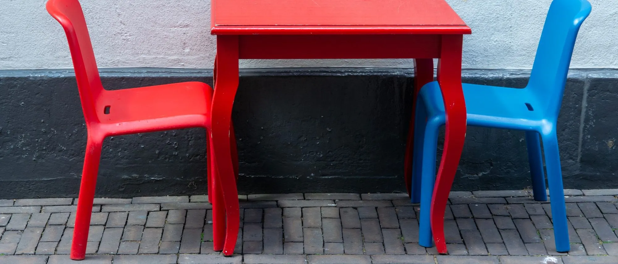 A red table with one red chair and one blue chair set out on a brick pavement.