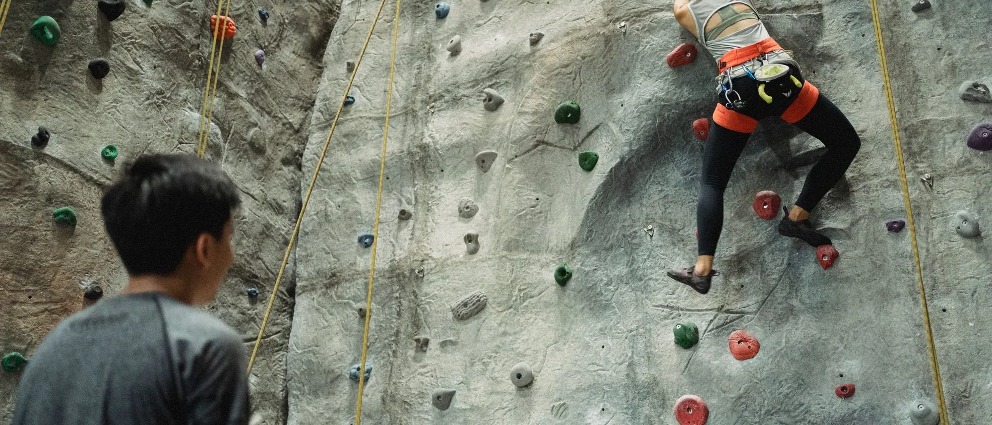 A climber on a grey indoor wall while a trainer watches from below, both seen from behind.