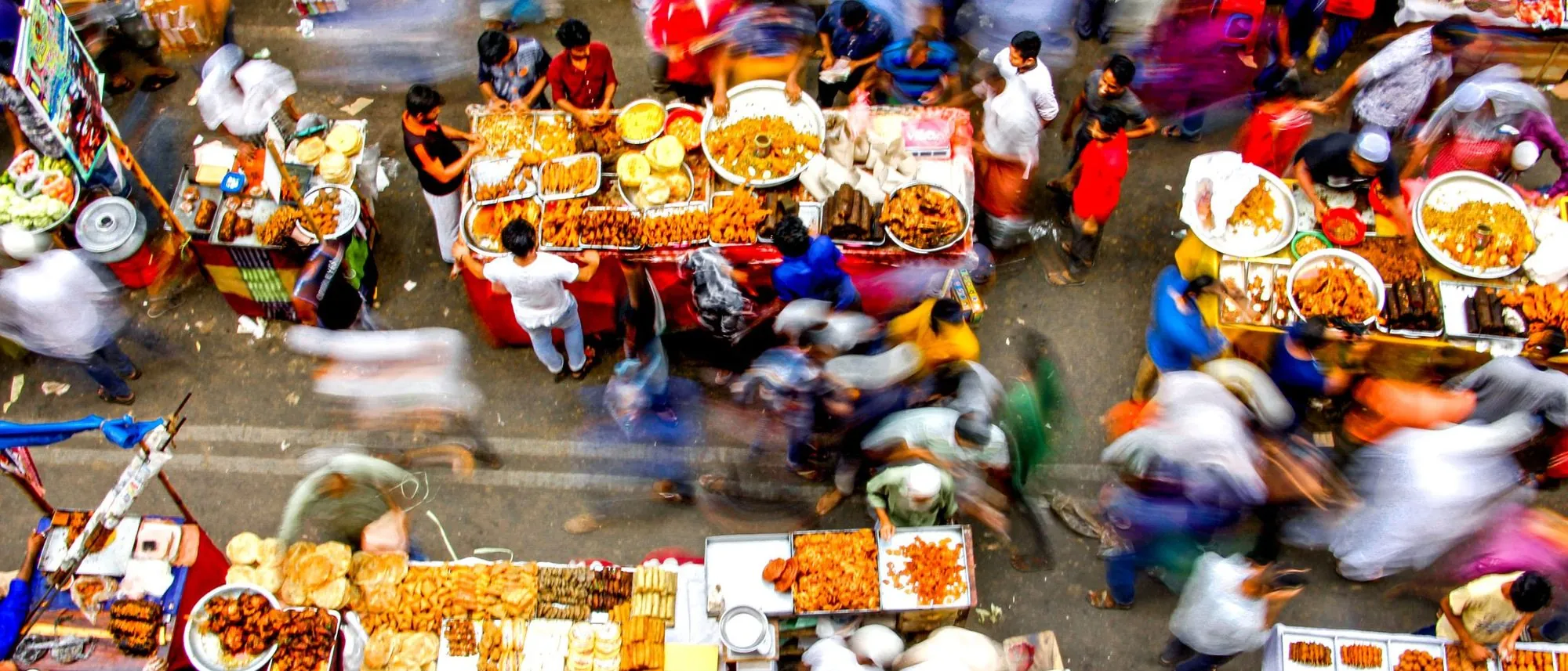 An overhead view of a crowded street market in Mumbai with food stalls and shoppers blurred by motion.
