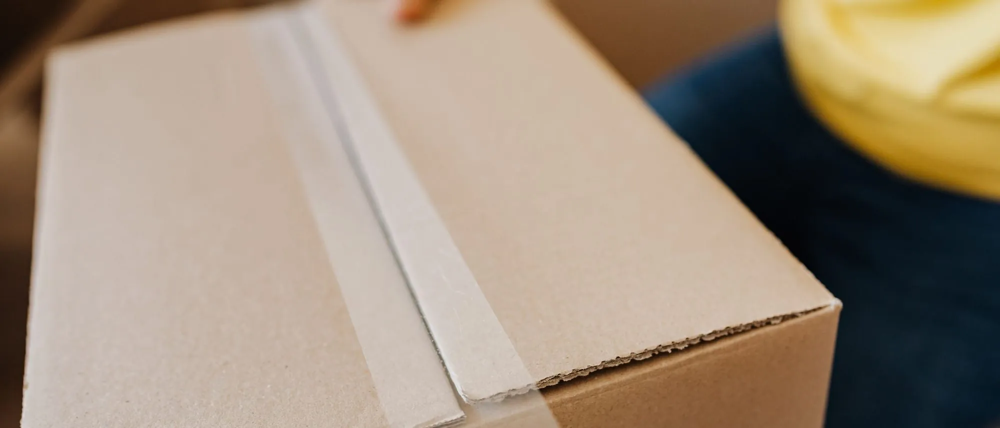 A person seals a cardboard box with clear tape, seen from the chest down.