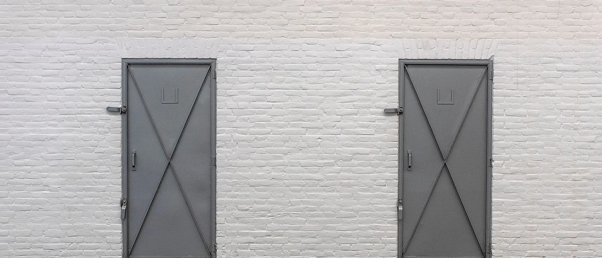 Two grey metal doors stand side by side in a white brick wall.