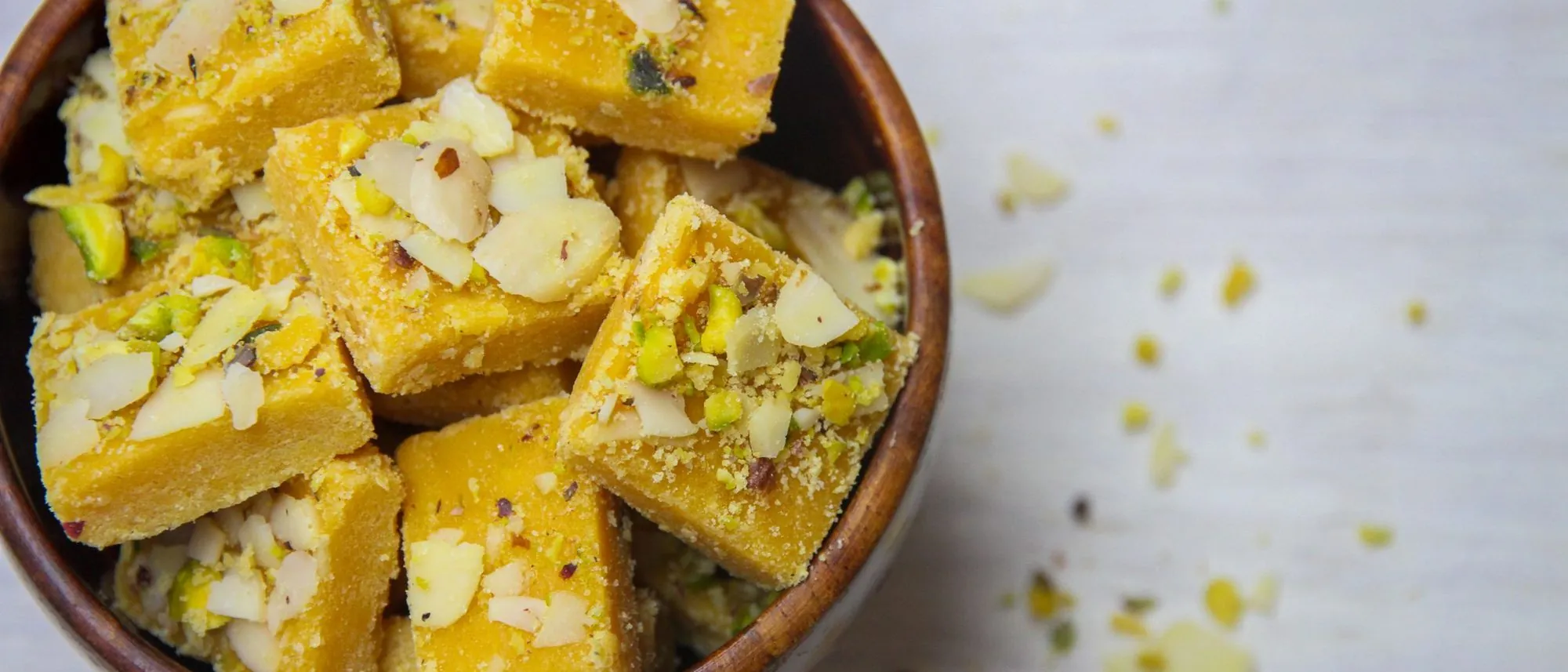 Pieces of besan barfi topped with almonds and pistachios in a wooden bowl seen from above.
