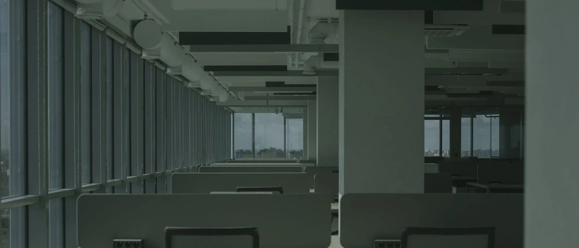 An empty office with rows of desks and large windows letting in daylight.
