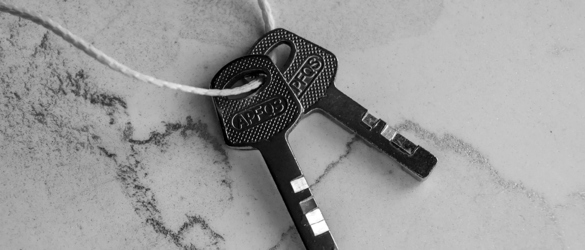 Two keys tied together with string on a textured marble surface, in black and white.