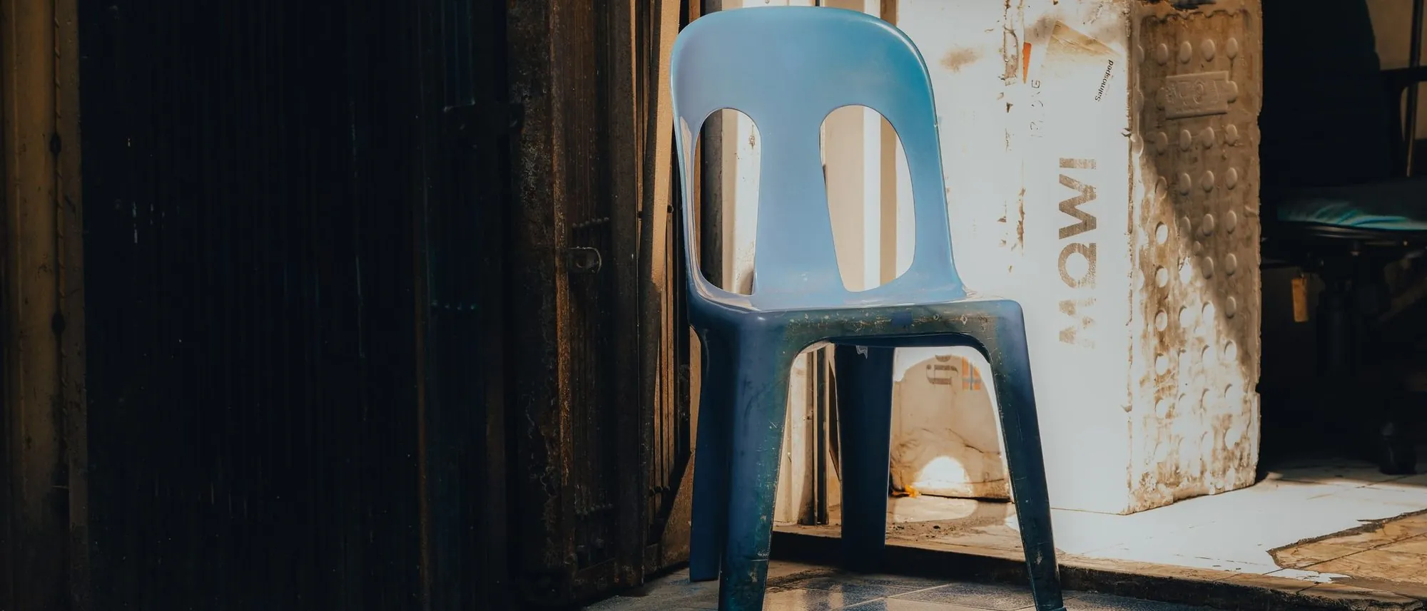 A single blue plastic chair in the corner of a sunlit room with worn floor tiles.