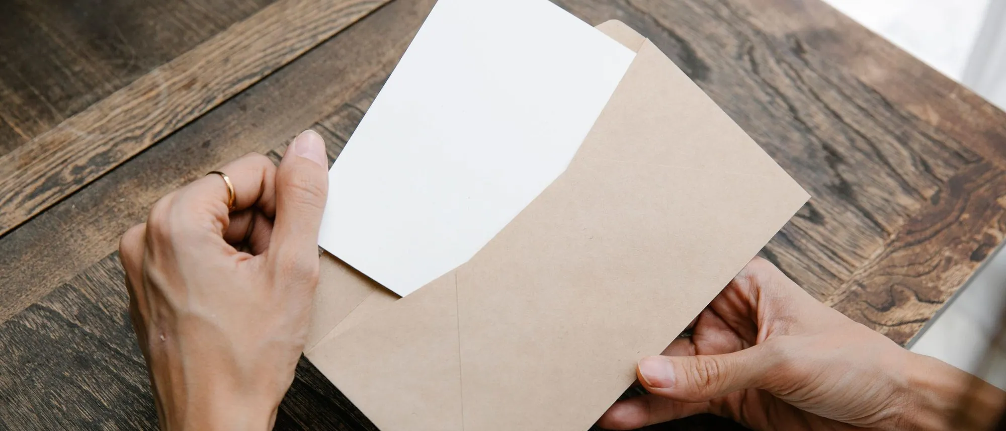 Seen from above two hands draw a blank sheet of paper out of a brown envelope on a wooden desk.