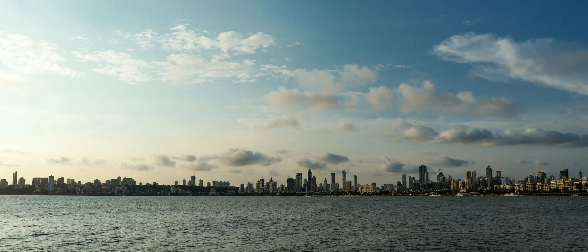 The Mumbai skyline stretches across the horizon beyond a calm sea under a cloudy sky.