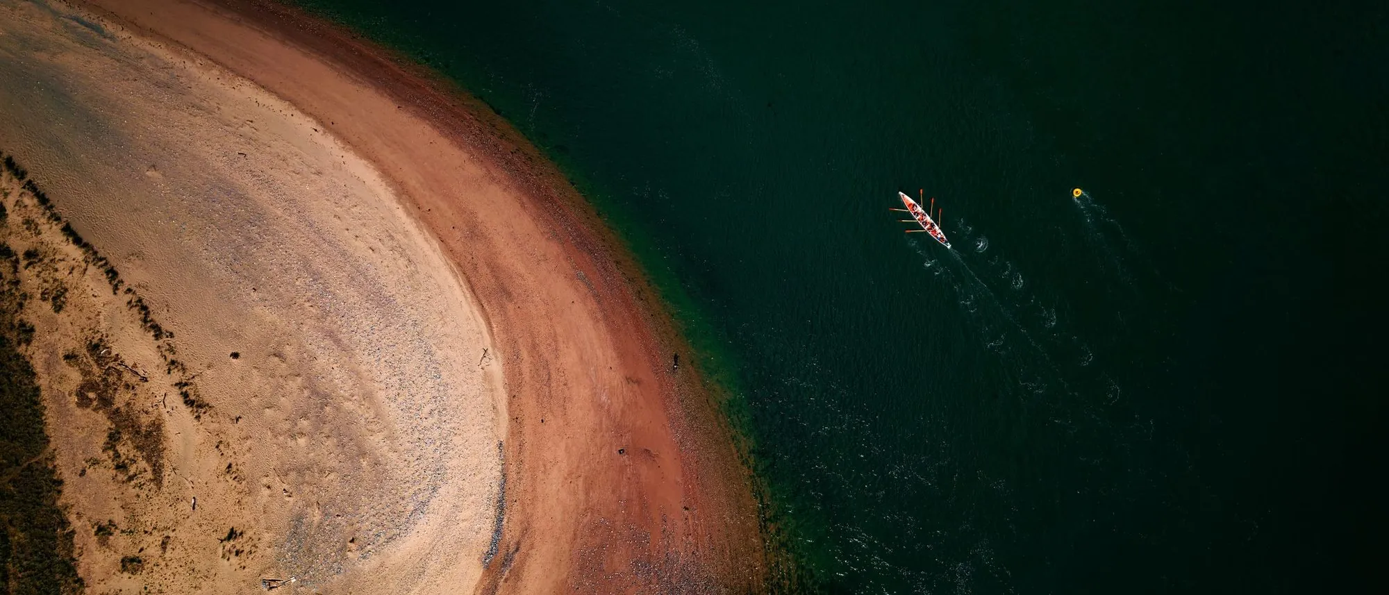 Rowers in a boat seen from above near a sandy shore and deep blue water.