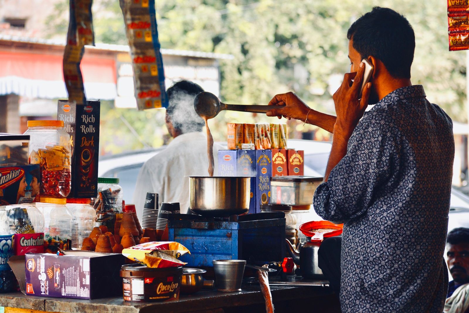 A street vendor talks on his phone while preparing tea at a market stall.