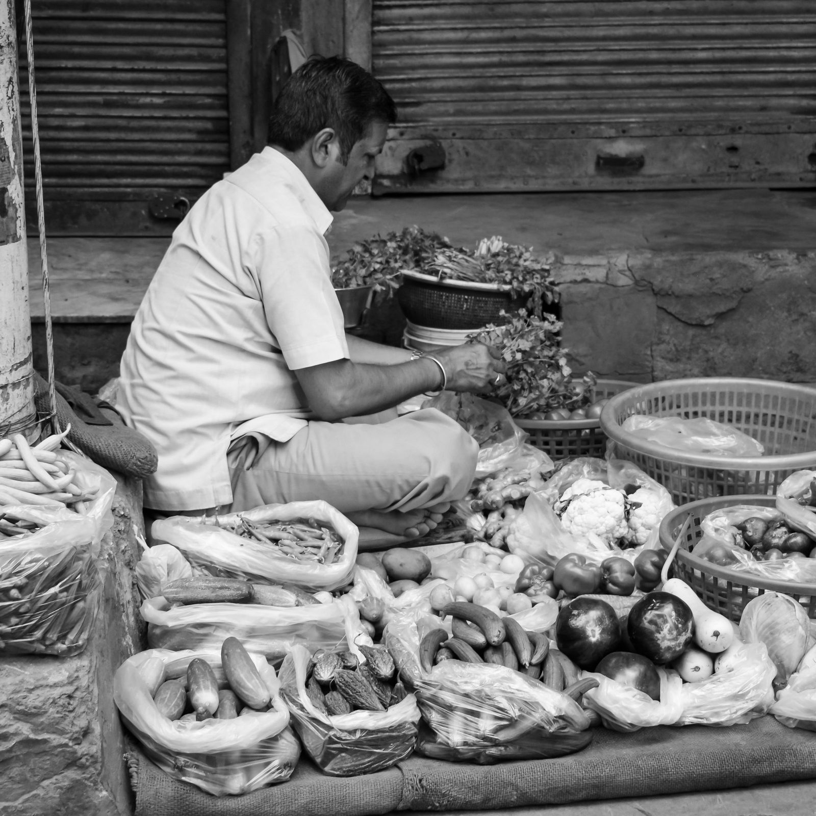 A vegetable seller surrounded by baskets of produce, in black and white.