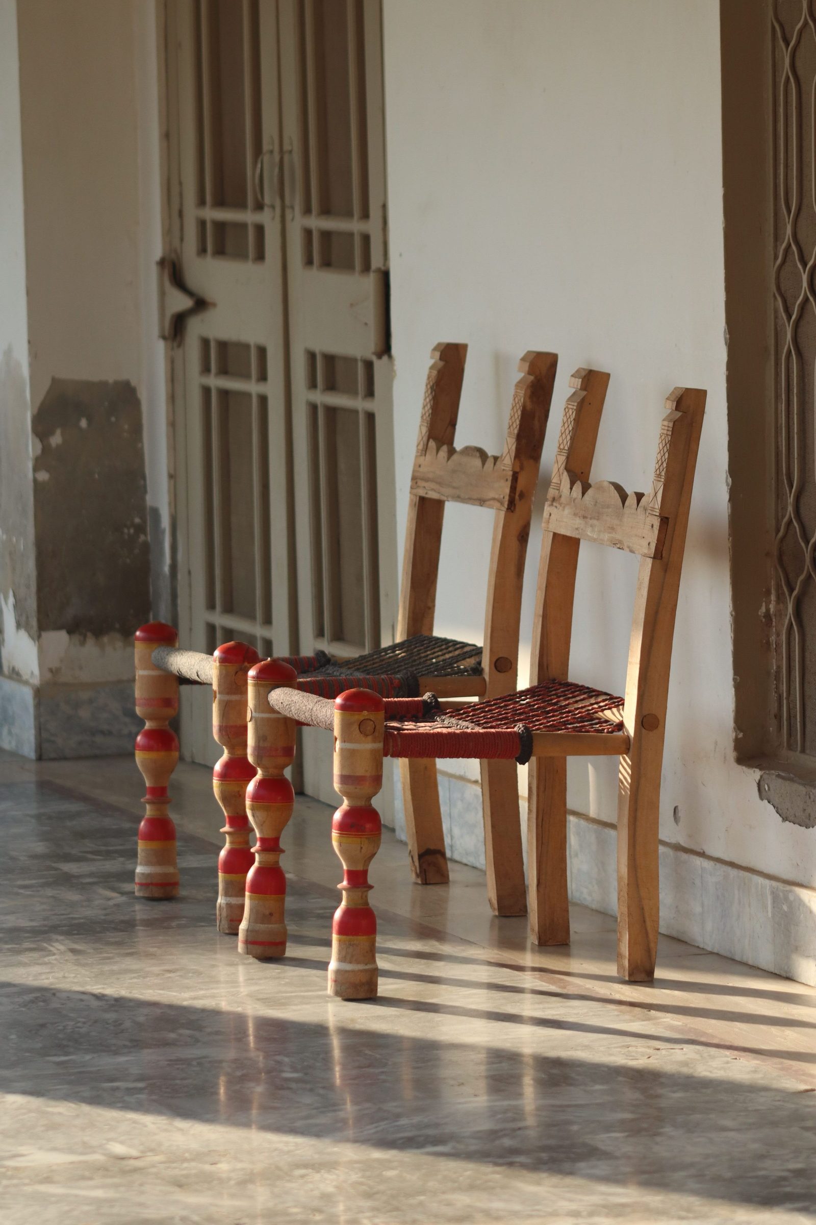 Two wooden chairs sit in a sunlit corridor of an Indian house.