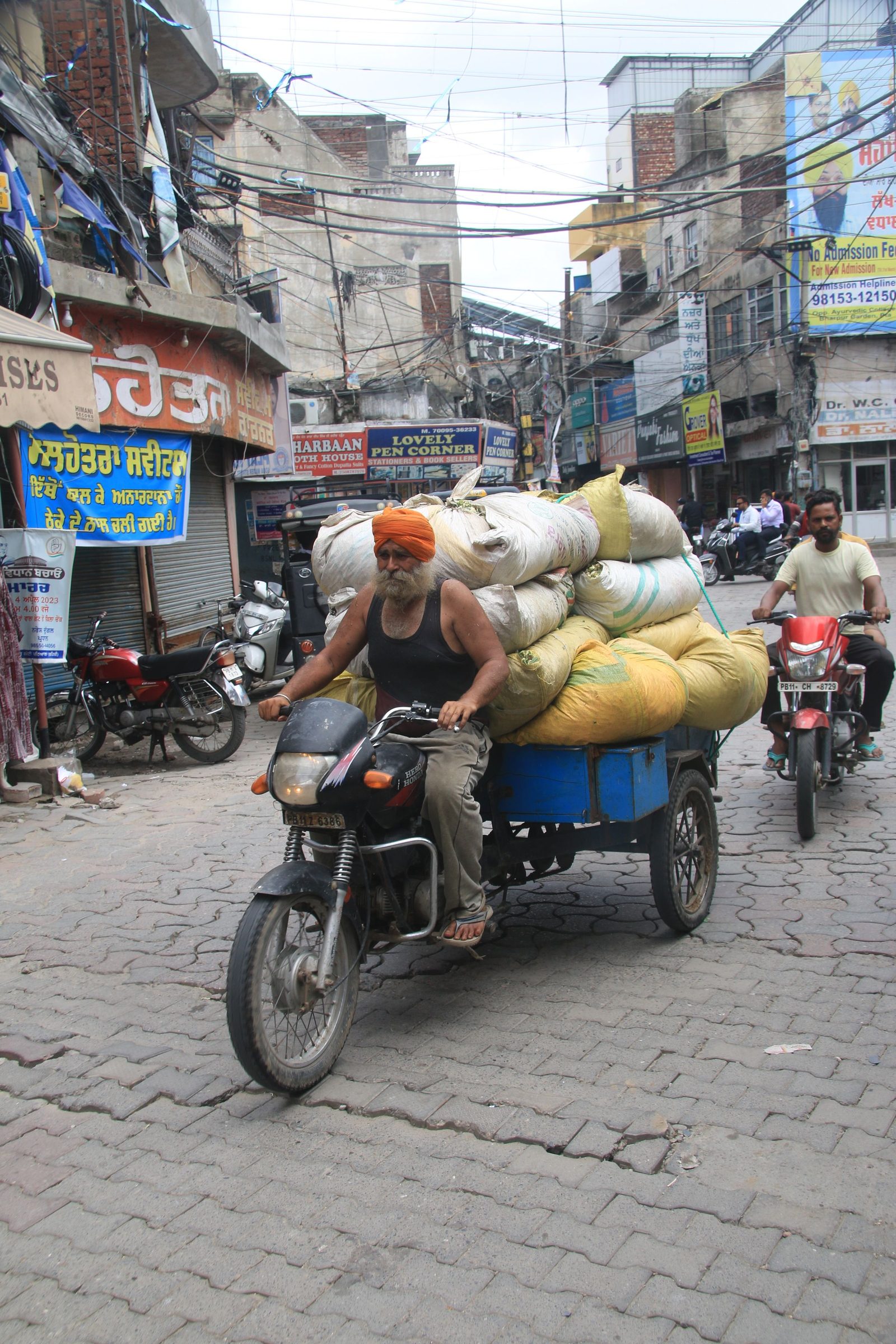 A man balances a heavy load of goods on a motorcycle in a crowded Indian street.