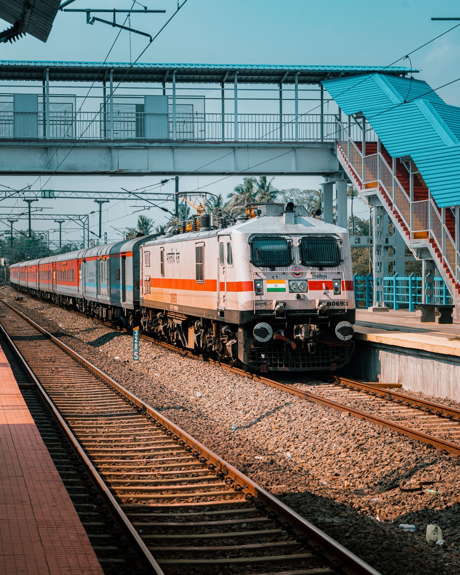 An Indian train passes under a station footbridge with tracks running into the distance.