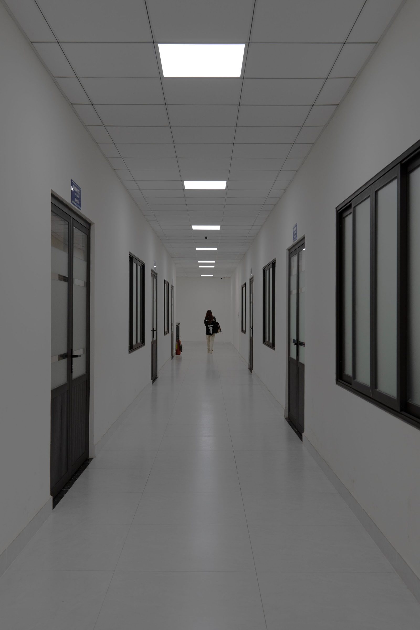 A figure walks away down a long white corridor lined with glass doors.