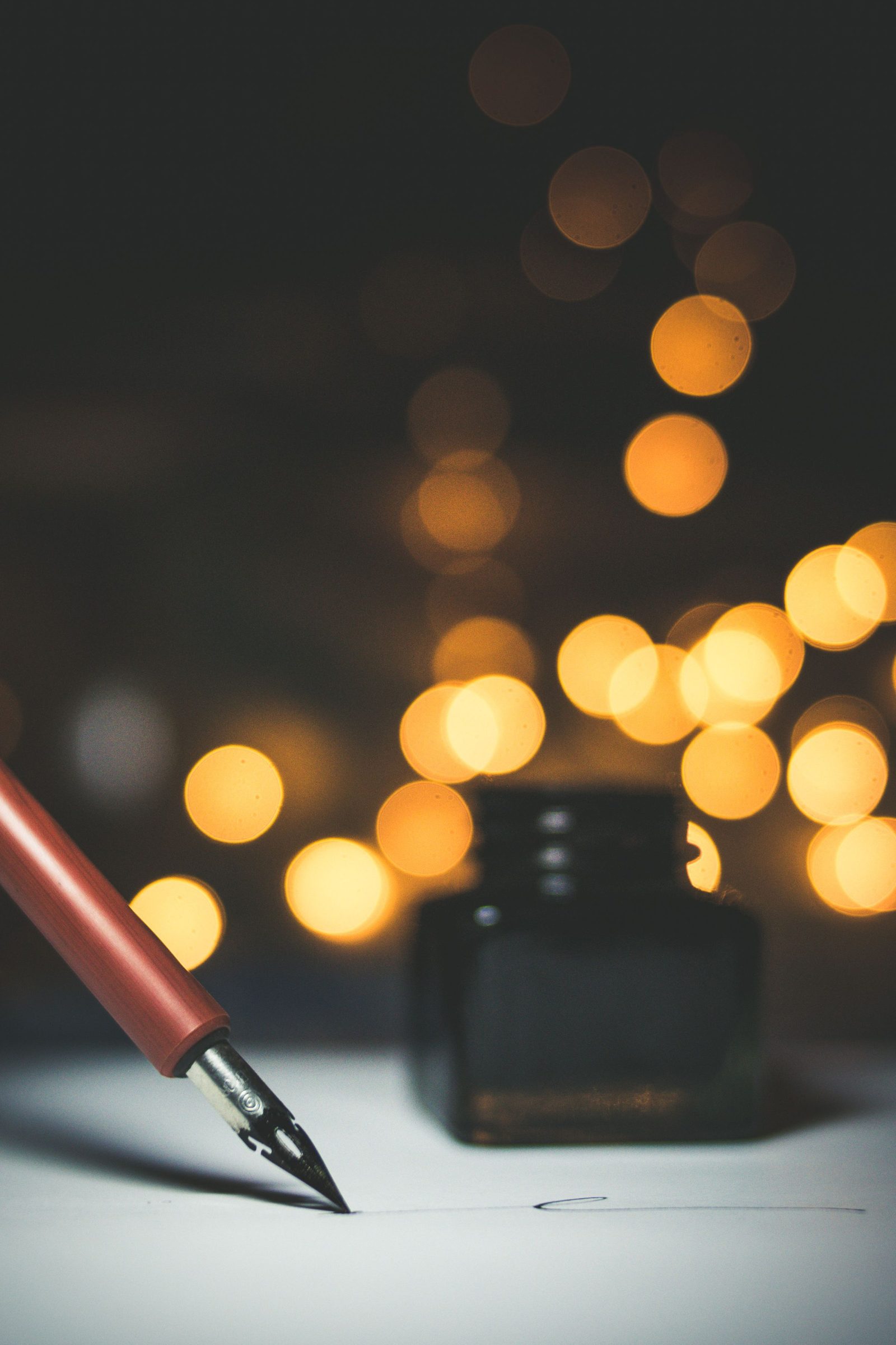 A fountain pen rests beside a bottle of ink against warm out-of-focus lights.