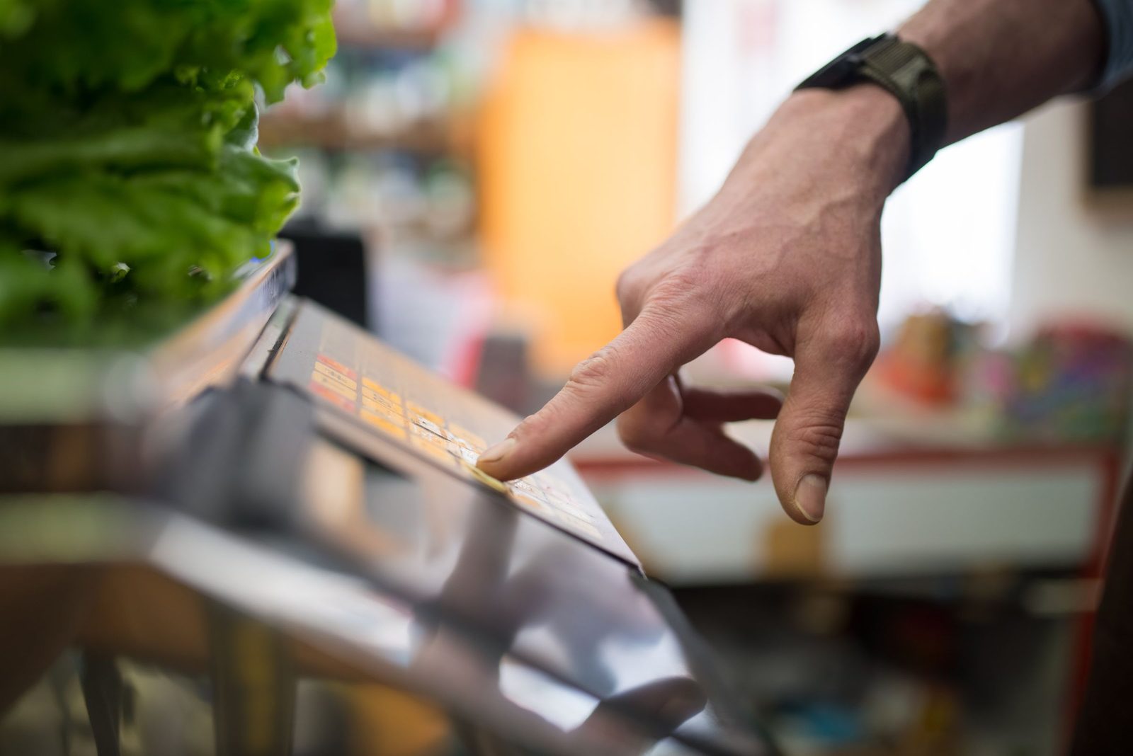 A hand presses the screen of a digital weighing scale in the produce aisle of a shop.