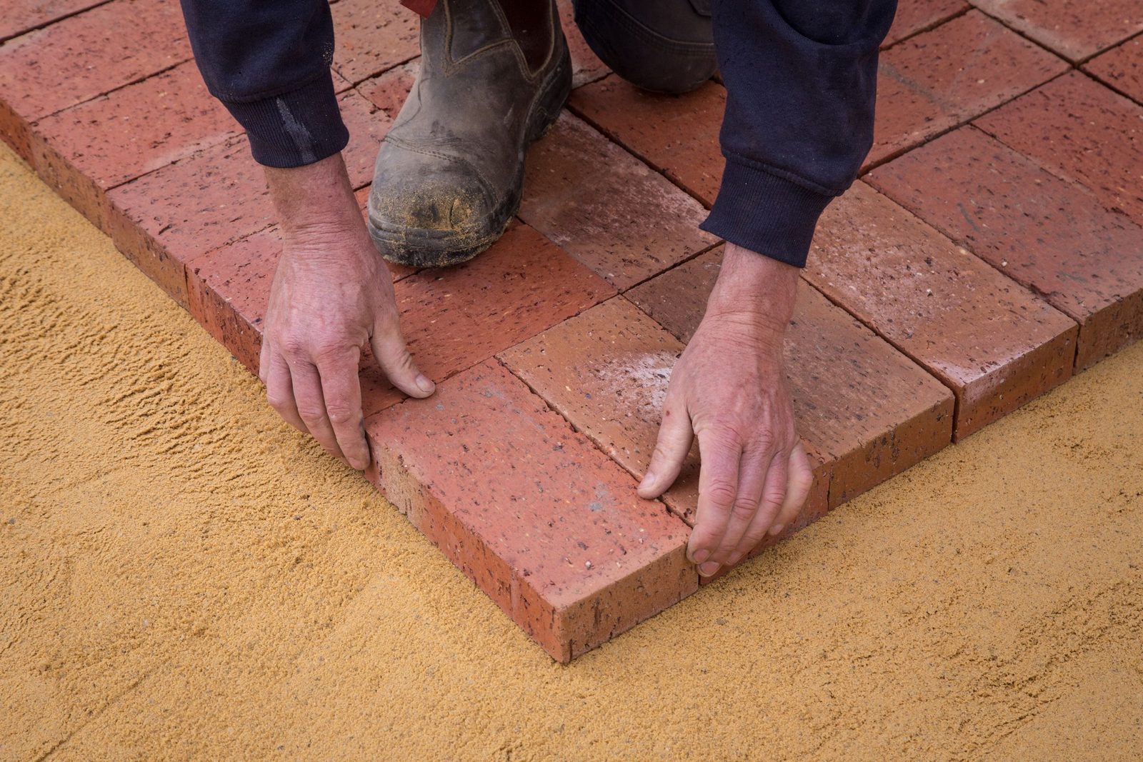 A worker aligns bricks into a square on sandy ground, seen from above.