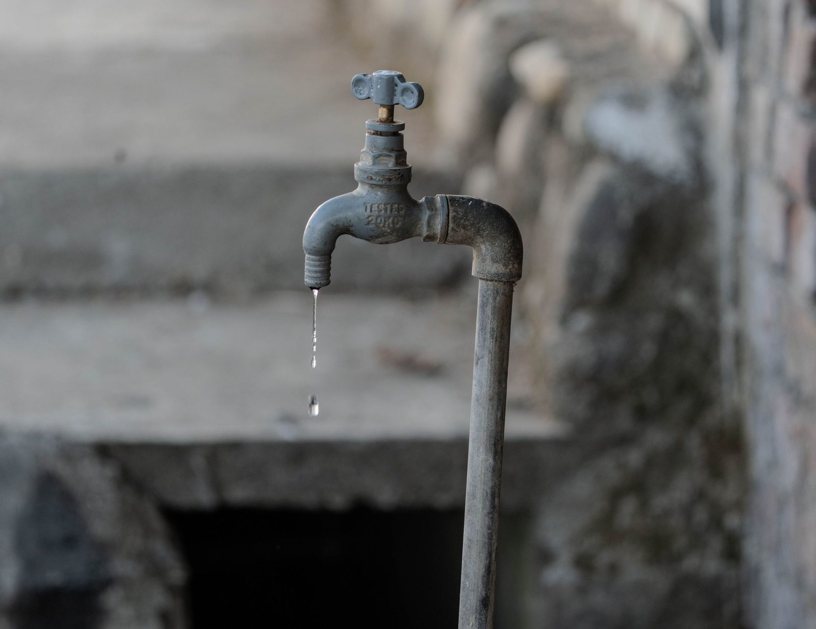 An old outdoor tap lets a single drop of water fall against a grey textured wall.