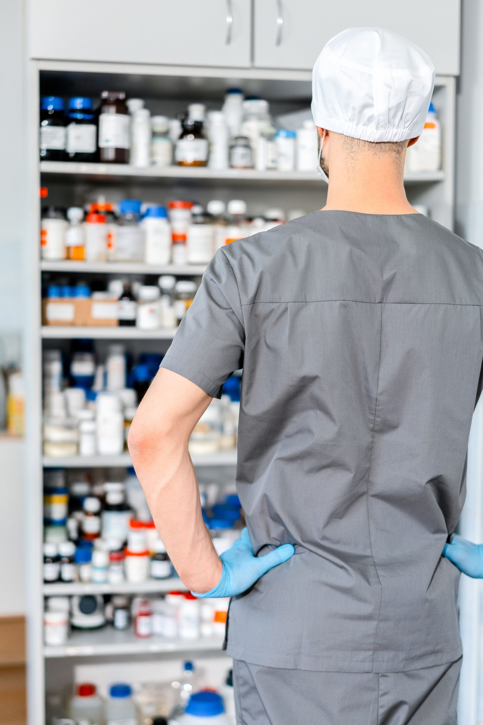 Seen from behind a pharmacist in a cap and gloves arranges shelves of medicine.