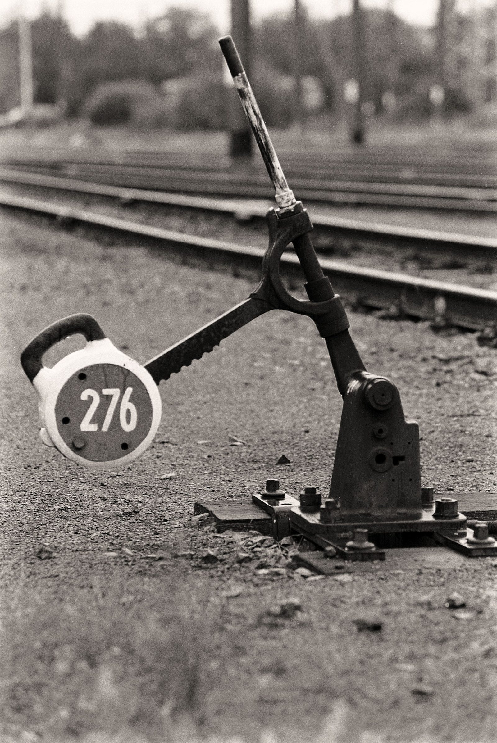 A black and white photograph of an old railway switch lever on gravel with tracks behind it.