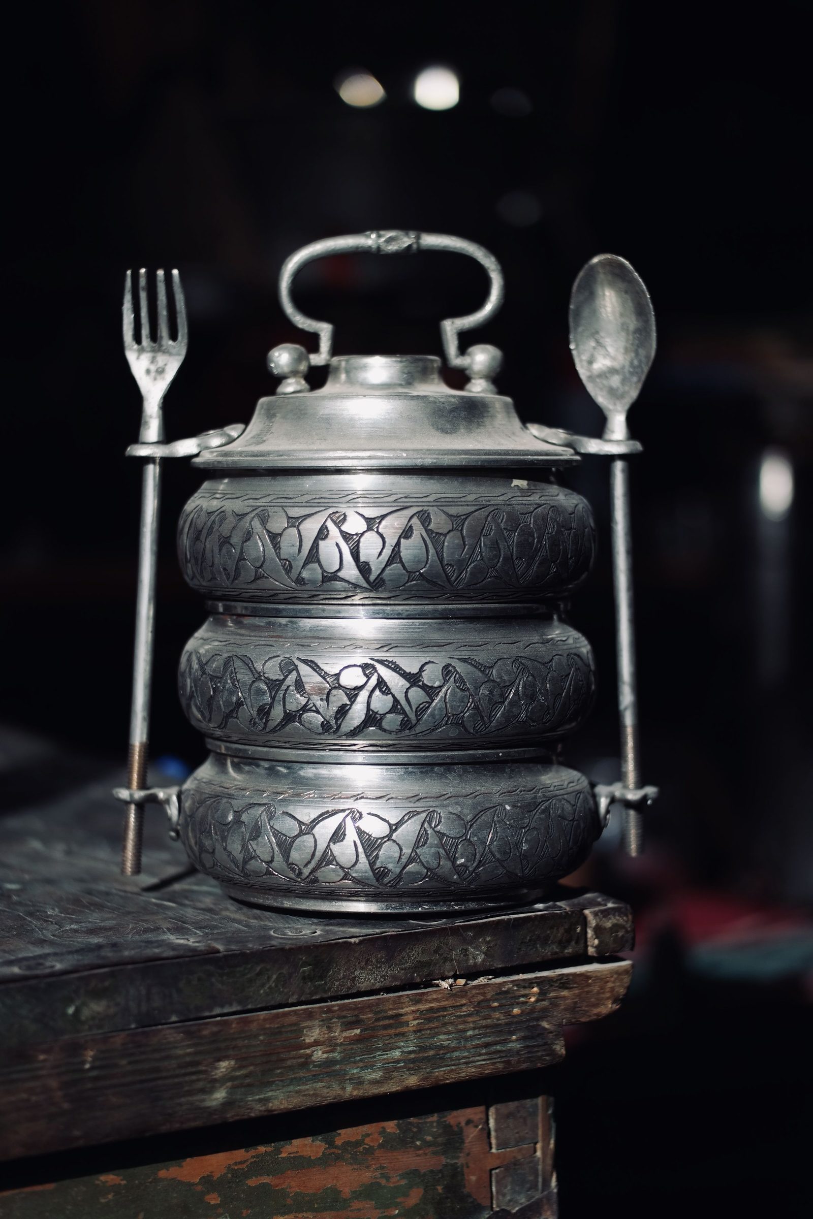 A vintage metal tiffin carrier with engraved tiers stands on a wooden surface.