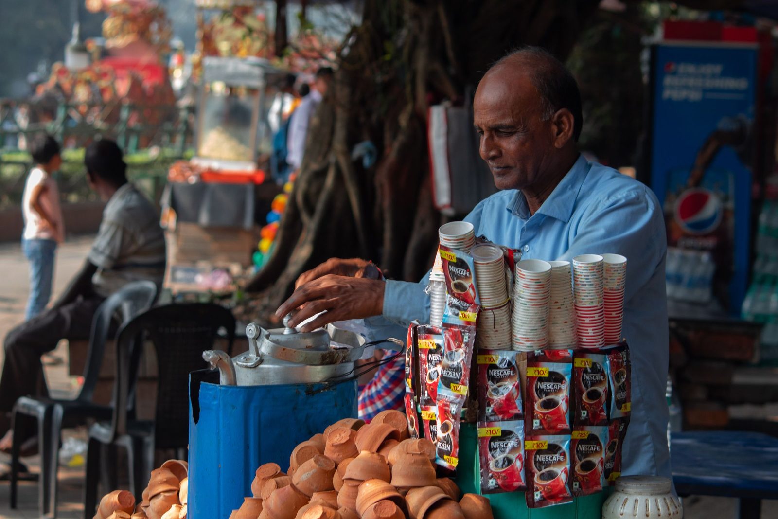 A vendor in a blue shirt serves chai in clay cups at a busy outdoor market stall.