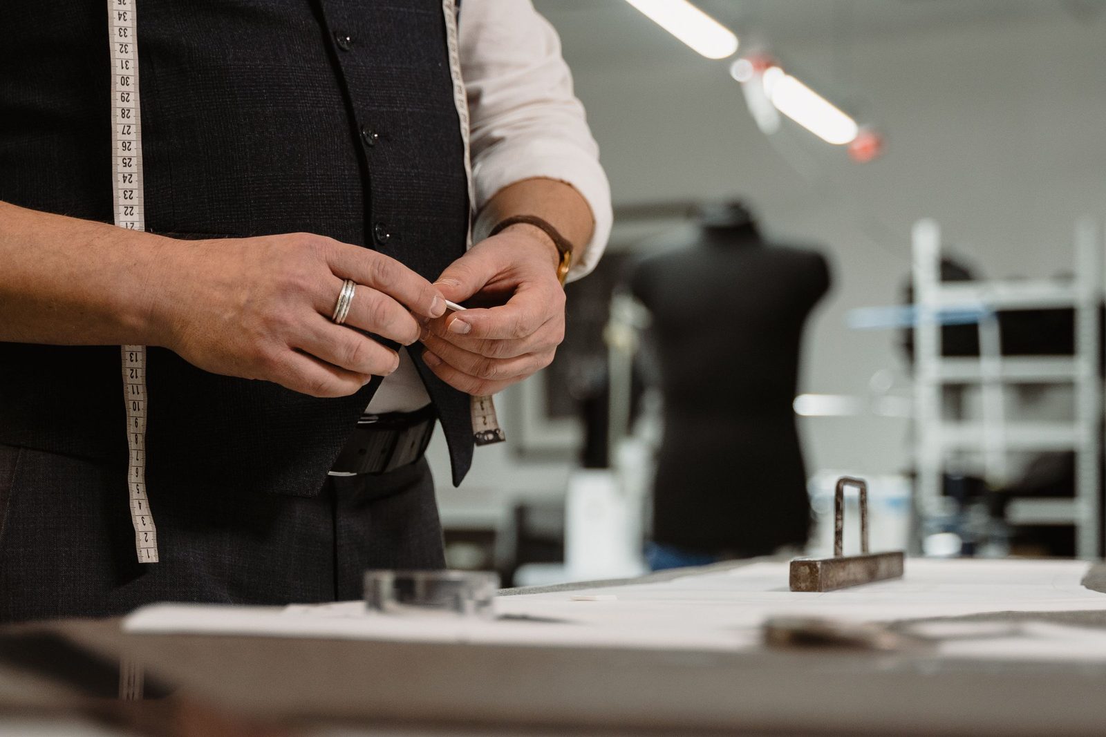 A tailor marks cloth with chalk beside a tape measure on the cutting table.