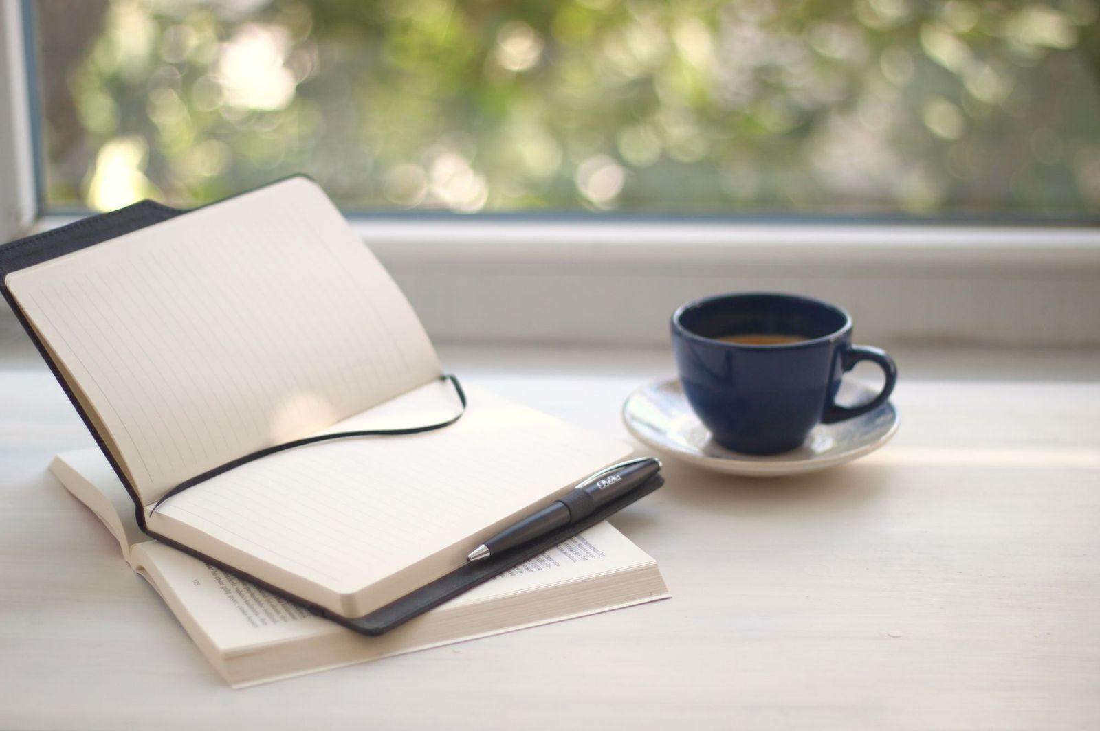 An open blank notebook with a pen beside a cup on a sunlit windowsill