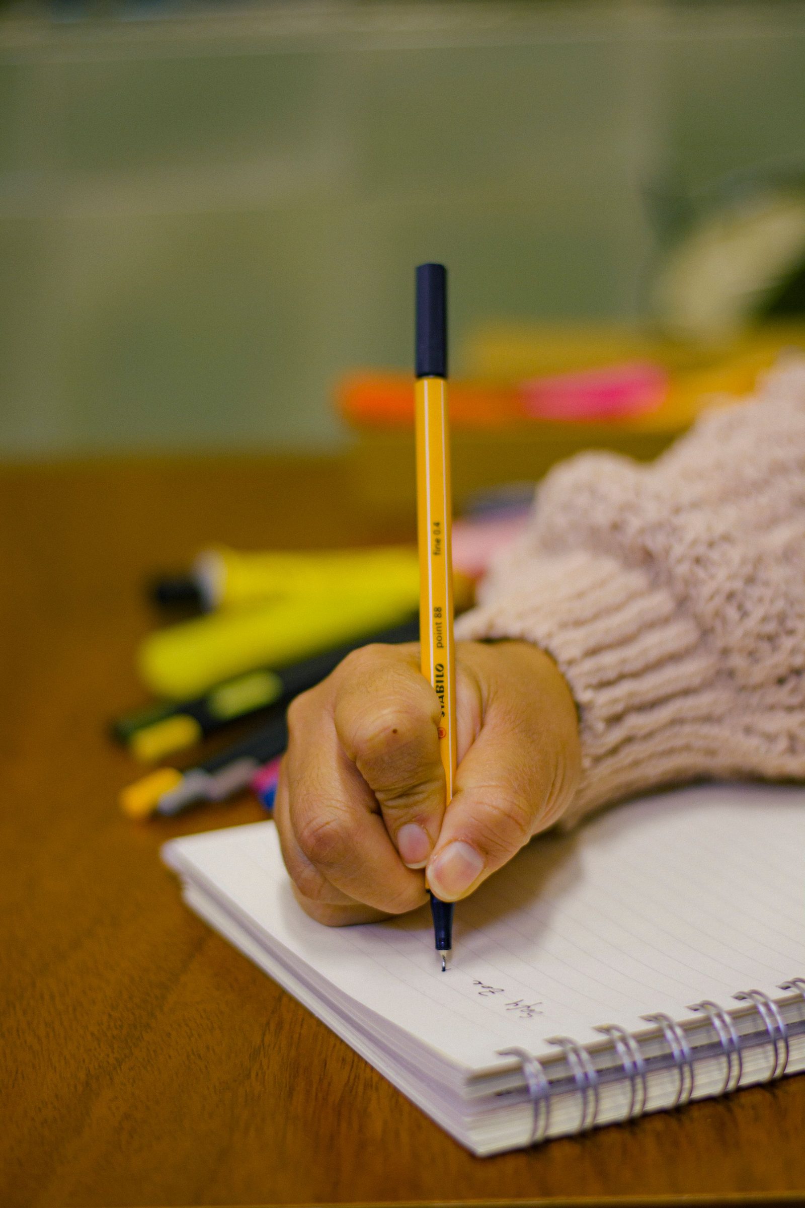 A hand writes in a notebook on a wooden table with pens scattered around.