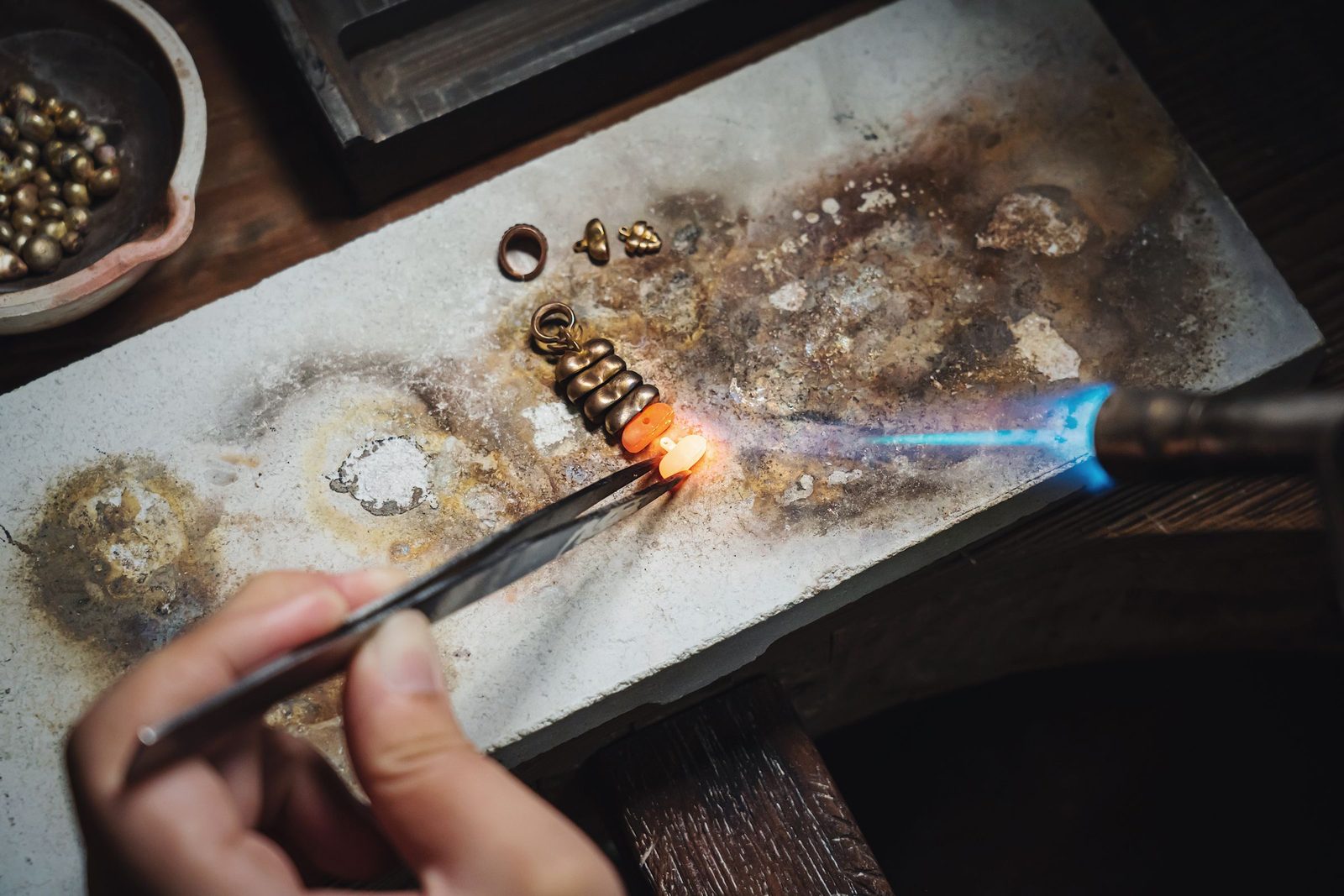A goldsmith’s hand holding a small gold piece in tweezers under a blue torch flame on a scorched work block