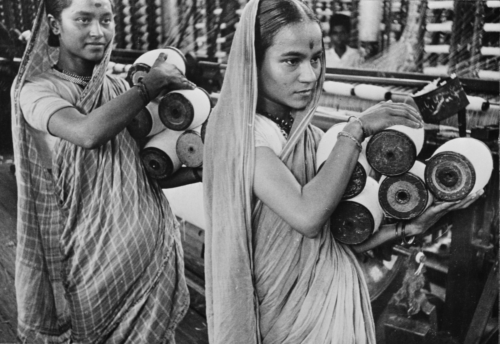 Two young women in saris carrying spools of thread in a textile mill