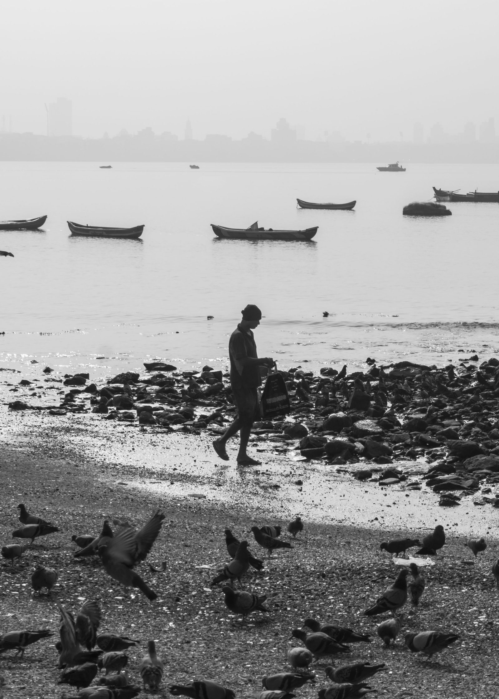 A man walks alone along a rocky Mumbai shore with boats and birds behind him in black and white.
