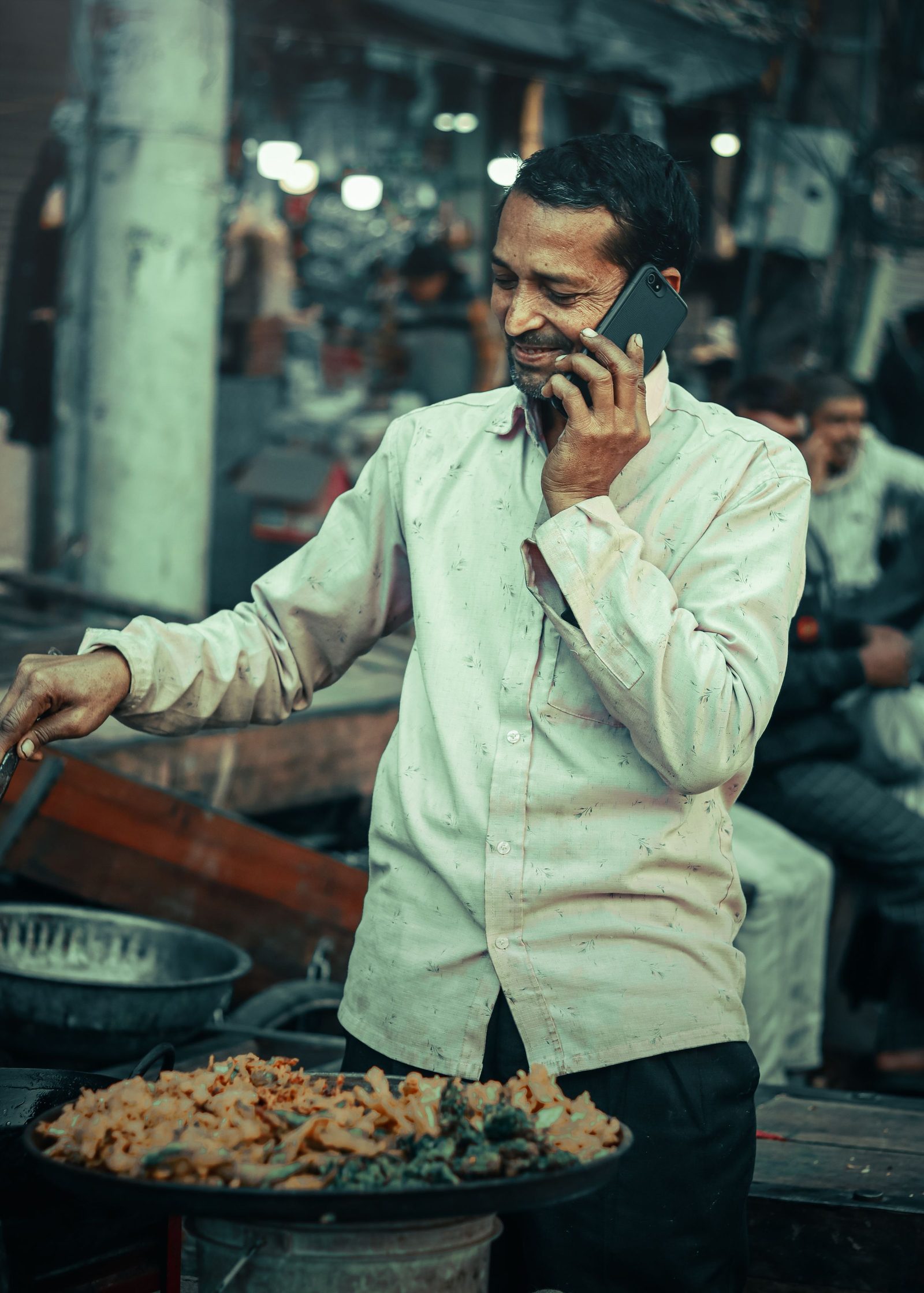 A street cook in a Delhi bazaar talks on his phone while tending a pan.