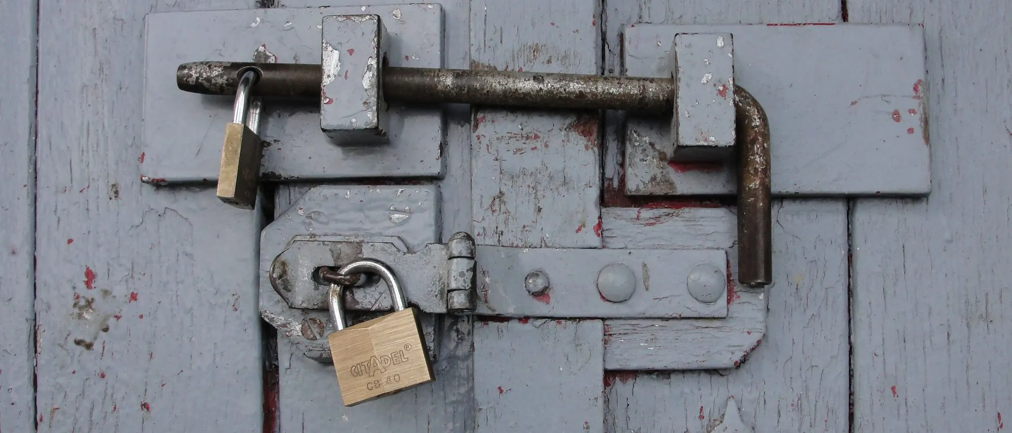 A weathered wooden door barred shut and secured with brass padlocks.