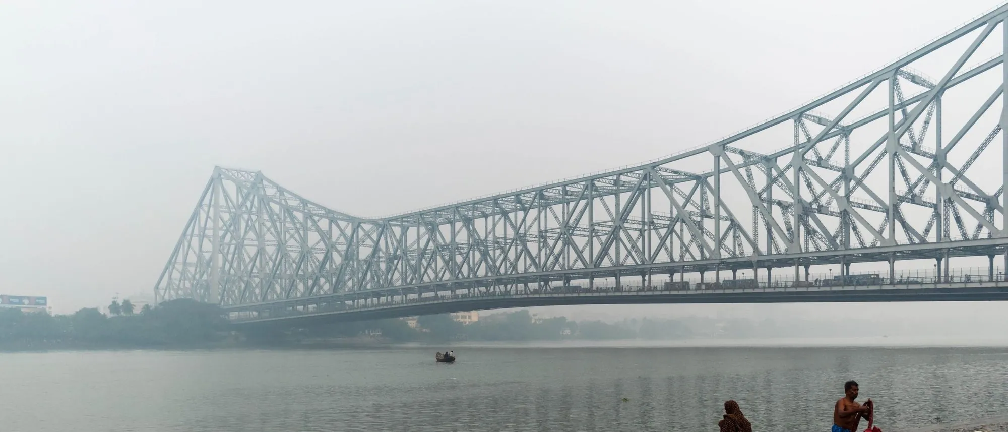 Howrah Bridge emerges from morning mist over the river with small figures on the bank.