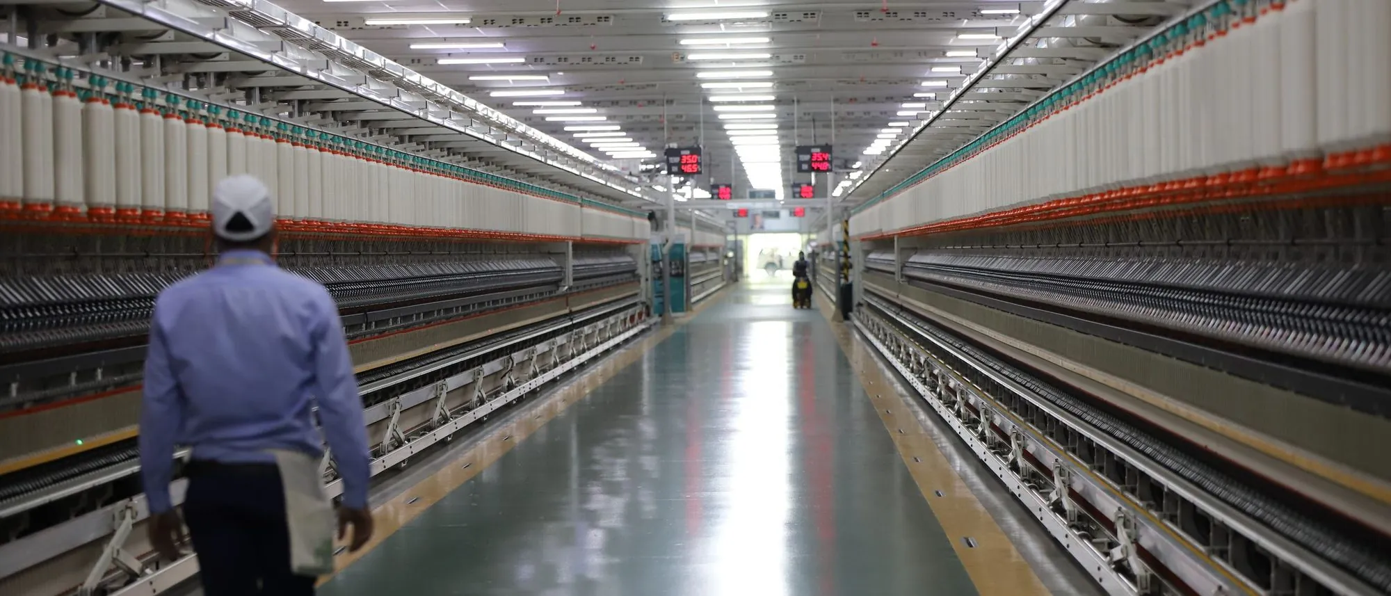 A long aisle between spinning machines in an Indian textile mill, with a single worker walking away down it.
