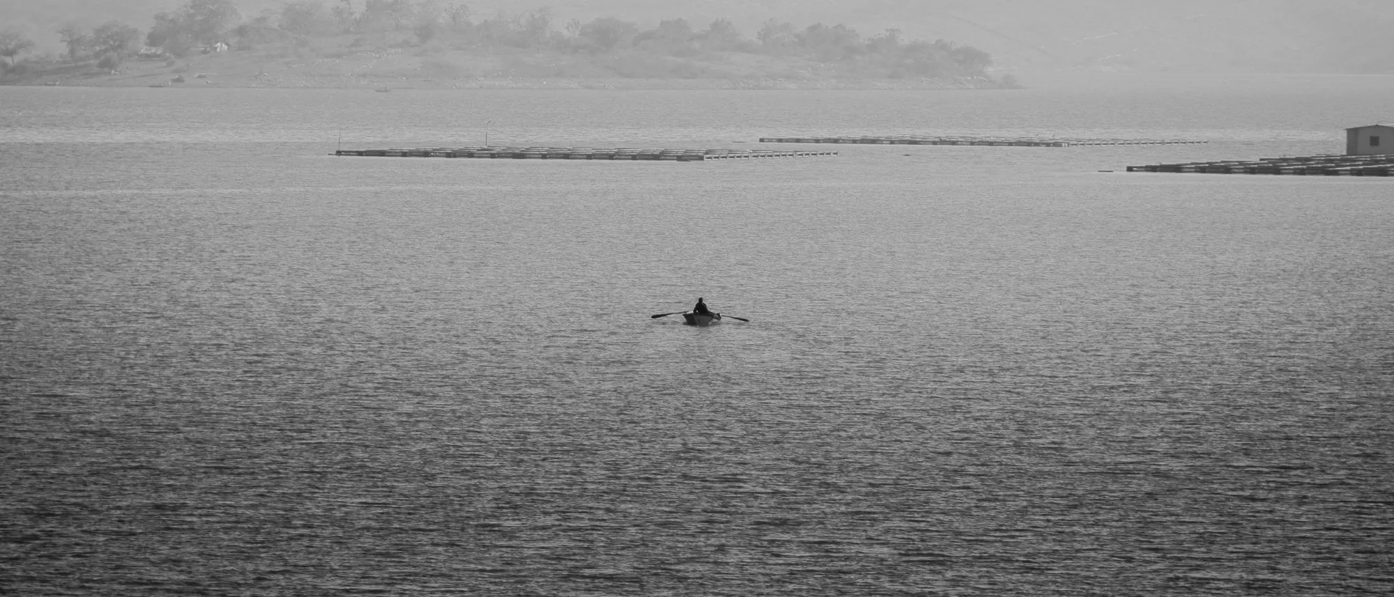 A single rowboat on a still lake in fog with hills fading behind it, in black and white.