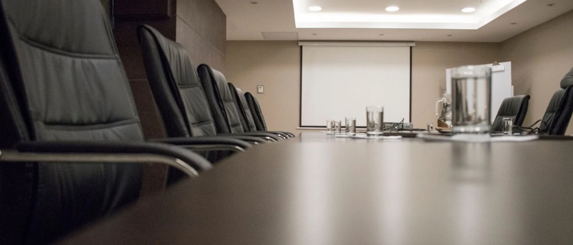 An empty conference room with black chairs around a long wooden table and glasses set out.