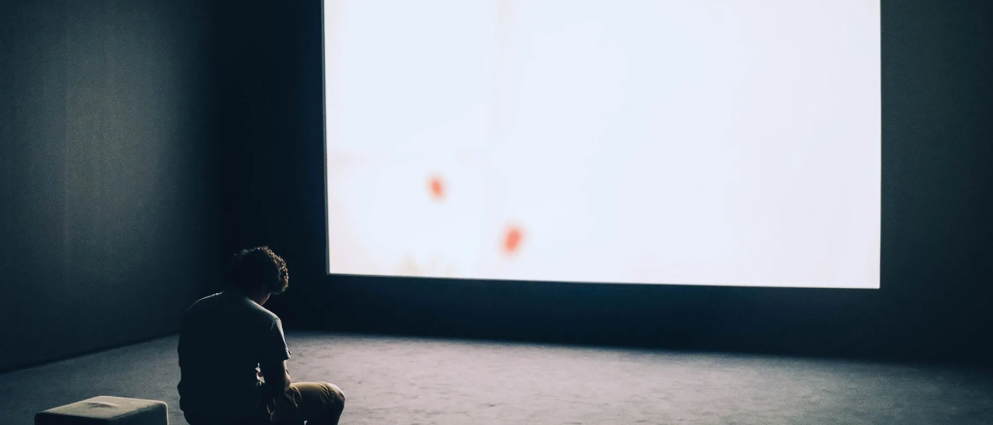 A man sits alone in a dark room facing a large blank white screen.