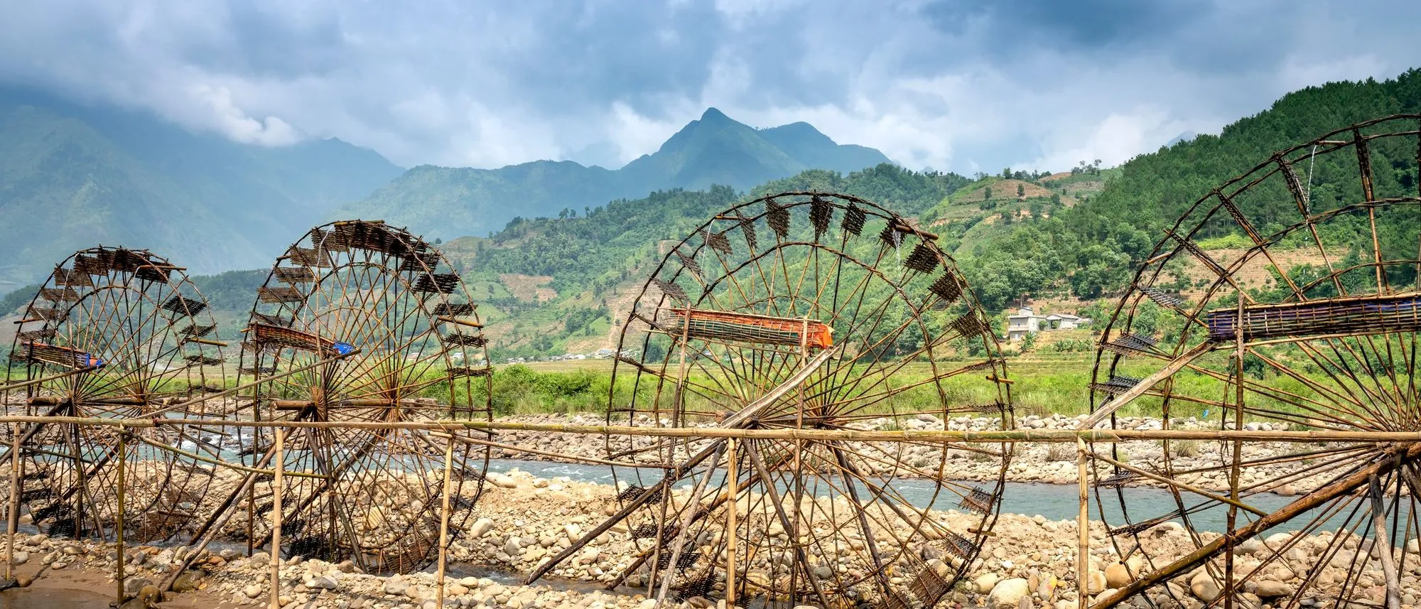 A row of wooden water wheels on a shallow river below misty green hills.