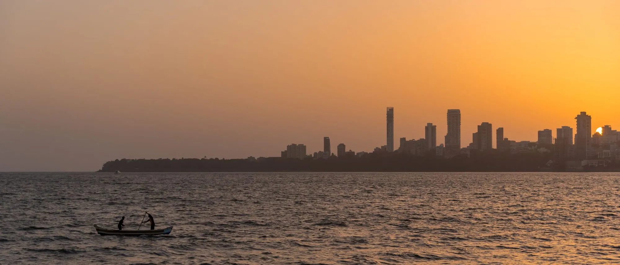 Two fishermen in silhouette on a small boat at sunset, with the Mumbai skyline behind them.