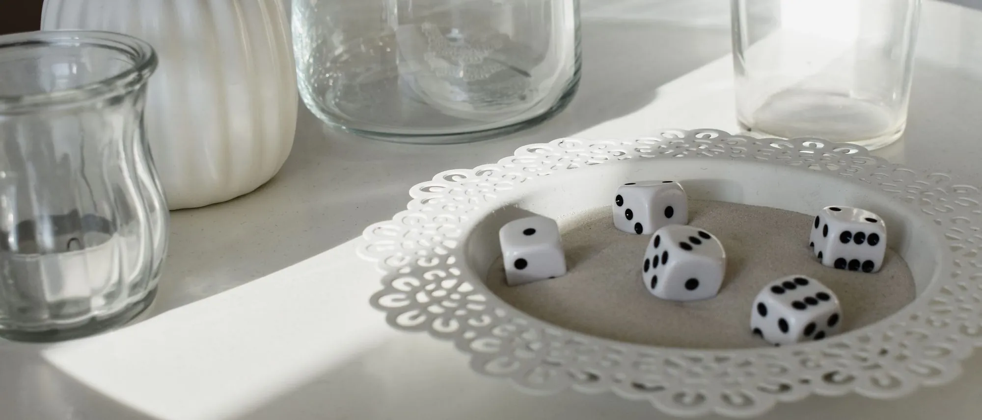 White and black dice sit among glassware and long shadows on a white table.