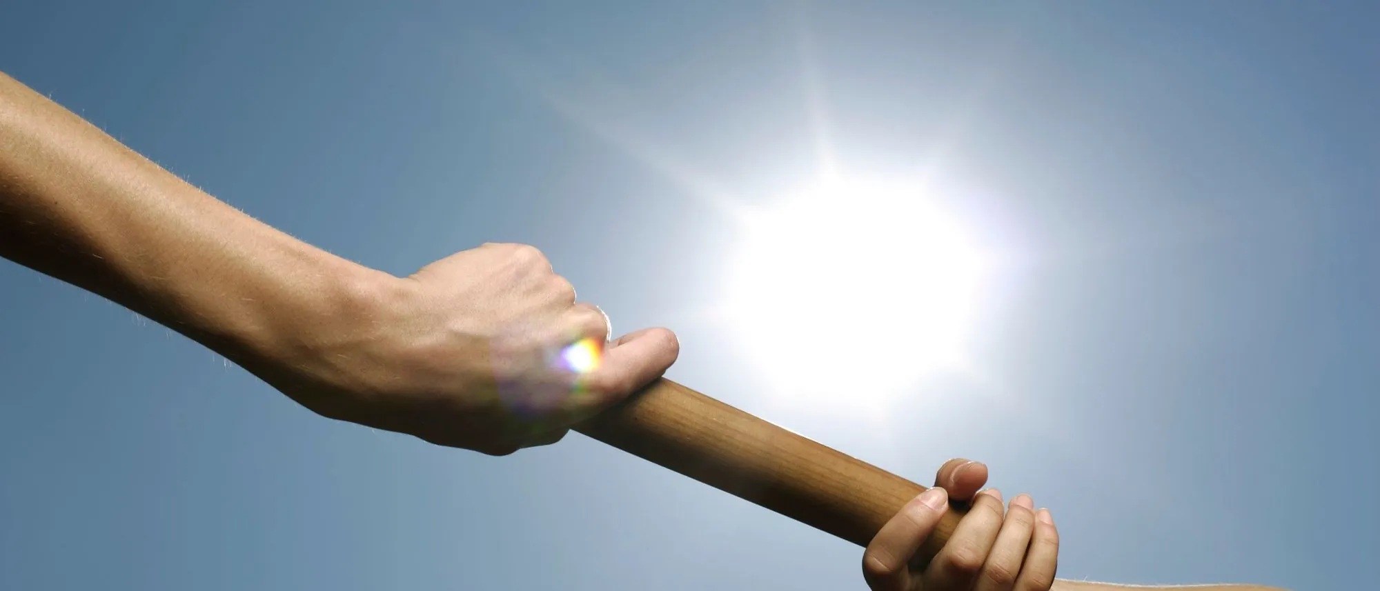 Two hands pass a relay baton against a bright sky.