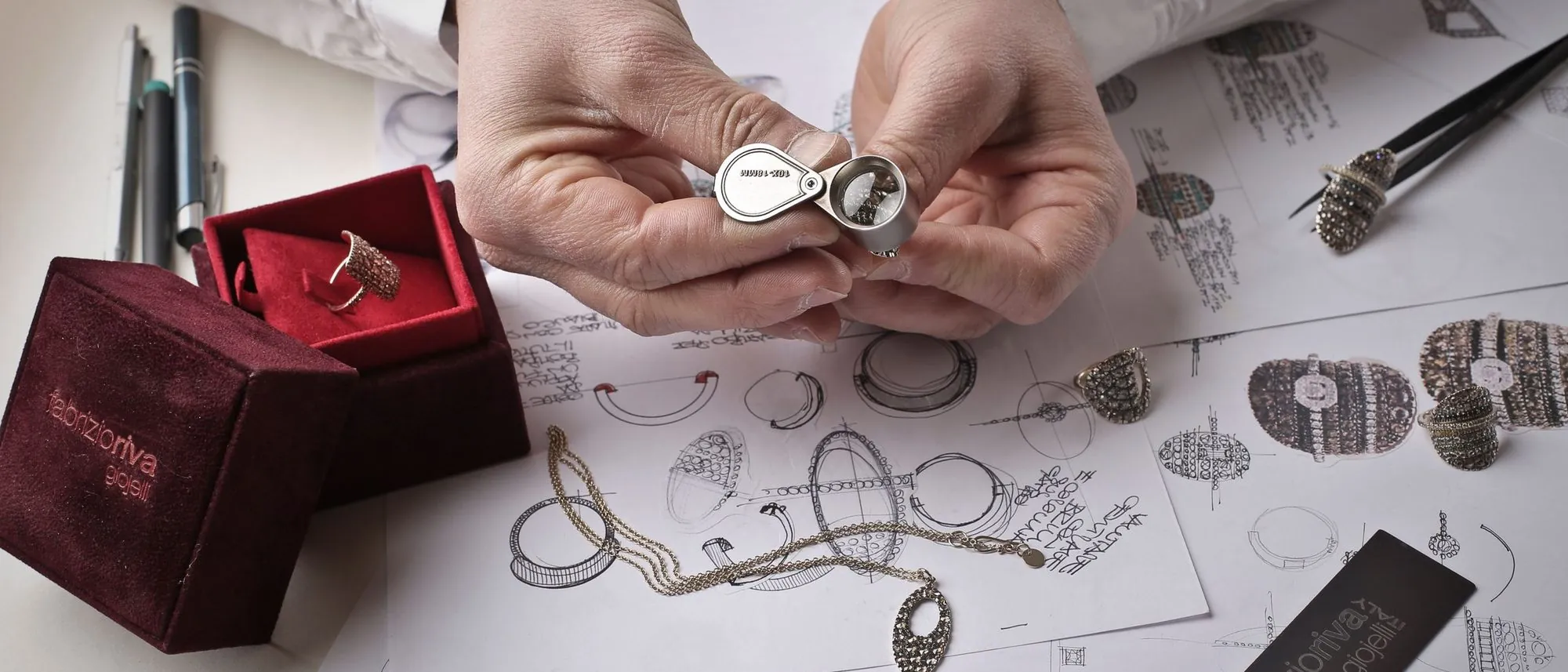 Two hands hold a ring under a jeweller’s loupe above a workbench scattered with sketches.