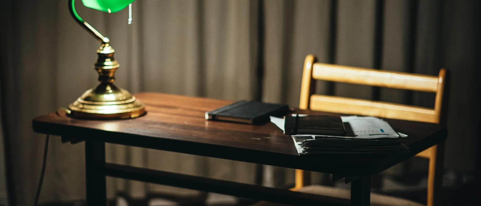 A dark room with a desk, an empty chair and one lit green banker's lamp